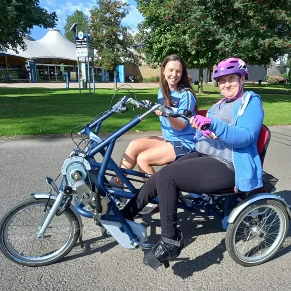 Two smiling women on a recumbent, tandem blue bike on a path surrounded by trees and Bells Sports centre in distance.