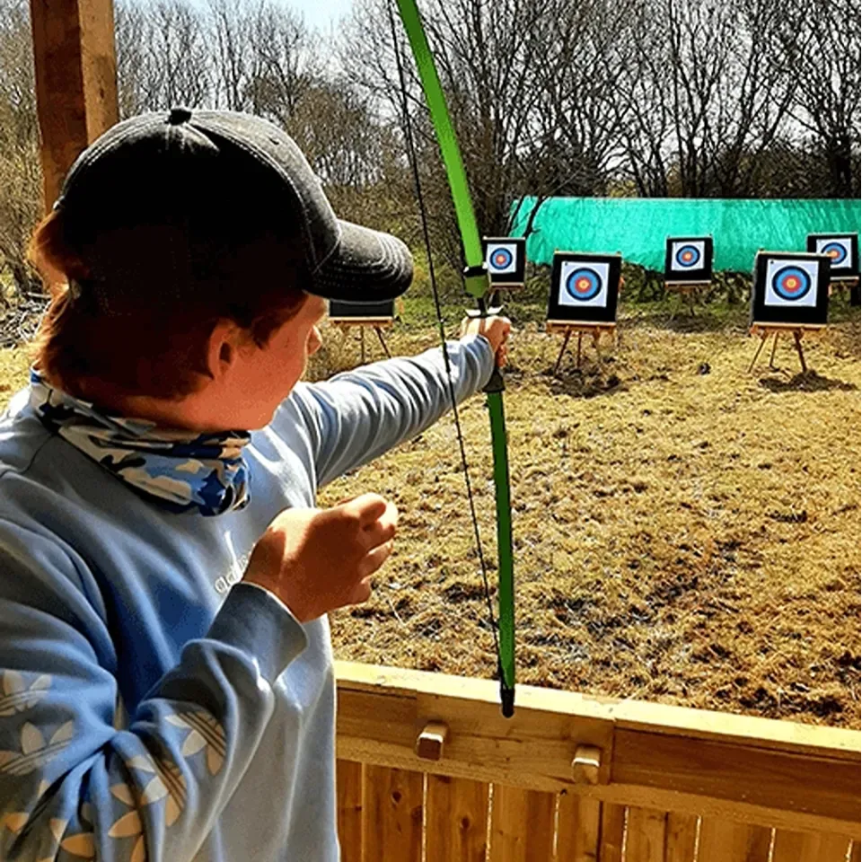 Young boy wearing a baseball cap in a wooden structure aiming a bow at an archery target