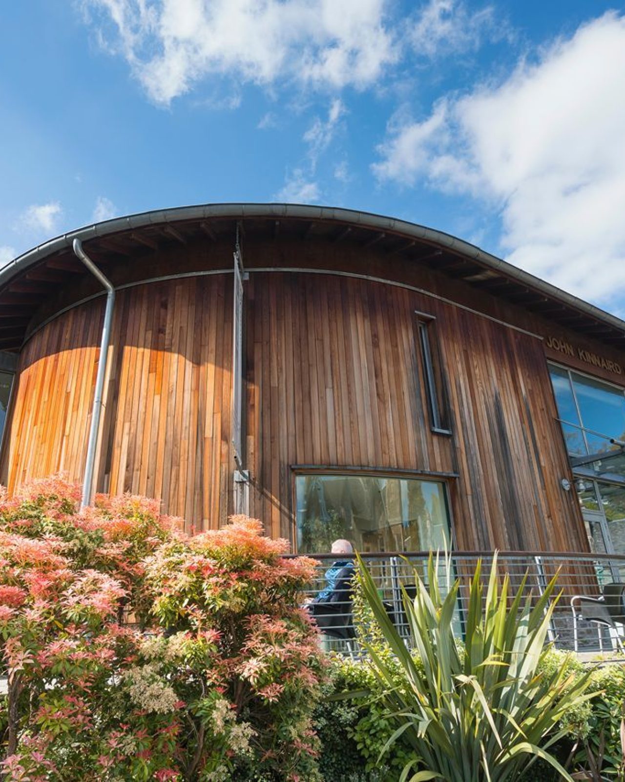 A modern wooden building surrounded by greenery and colourful flowers under a blue sky with clouds.