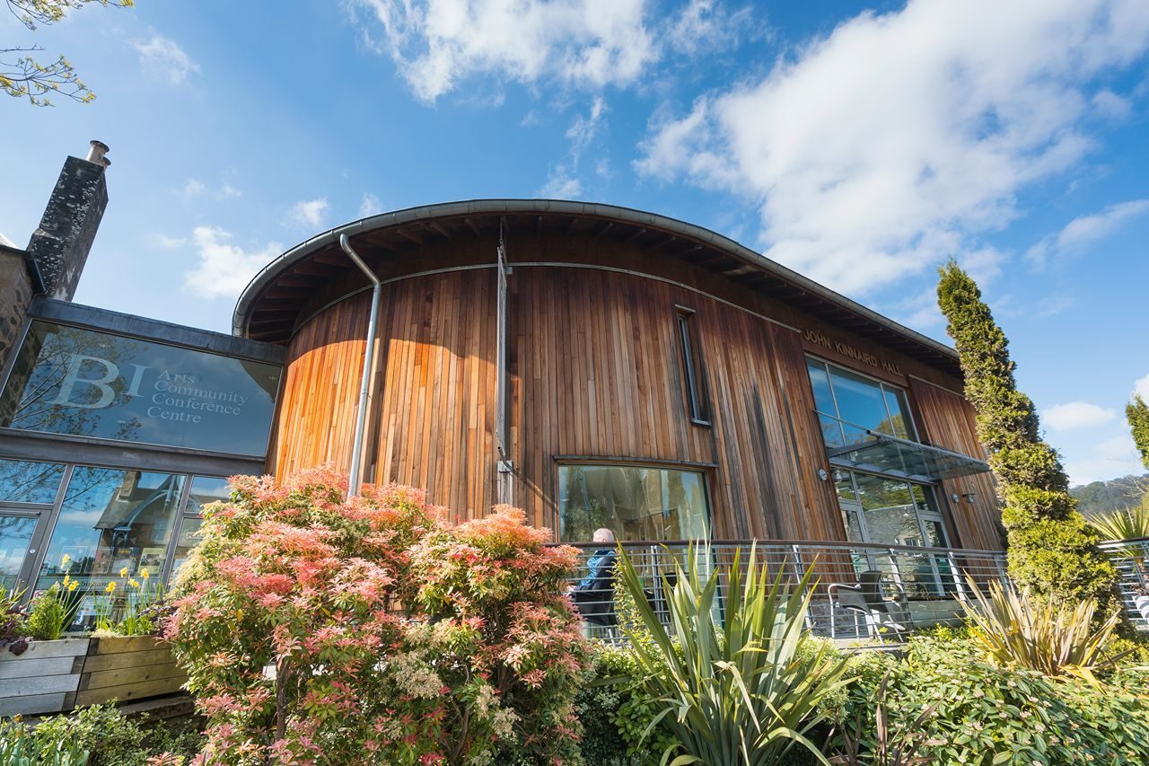 A modern wooden building surrounded by greenery and colourful flowers under a blue sky with clouds.