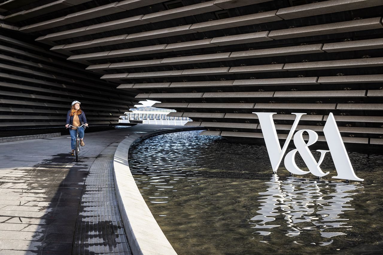 A woman riding a bike along a waterway, passing a sign that reads "V&A."