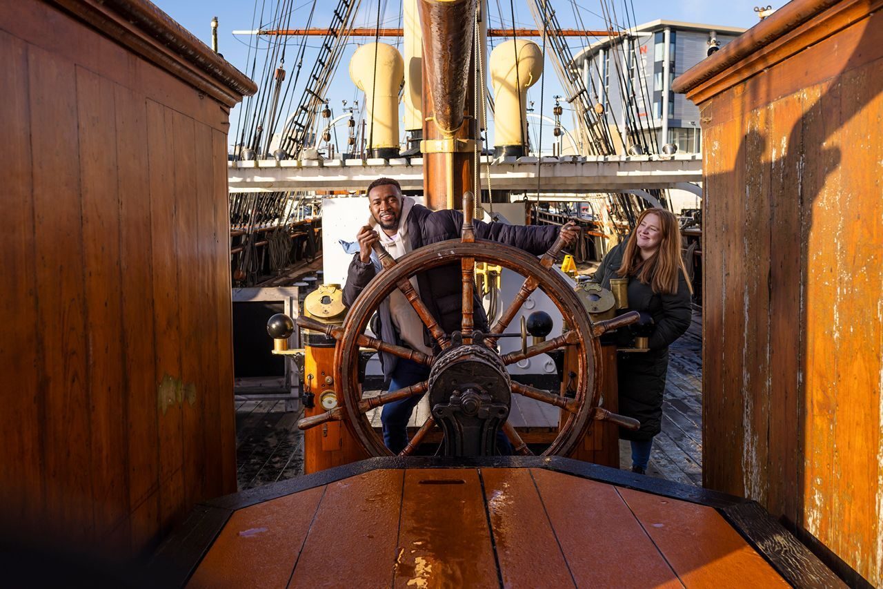 Two people enjoying a day on a historic ship, smiling at the helm with wooden accents and nautical elements in the background.