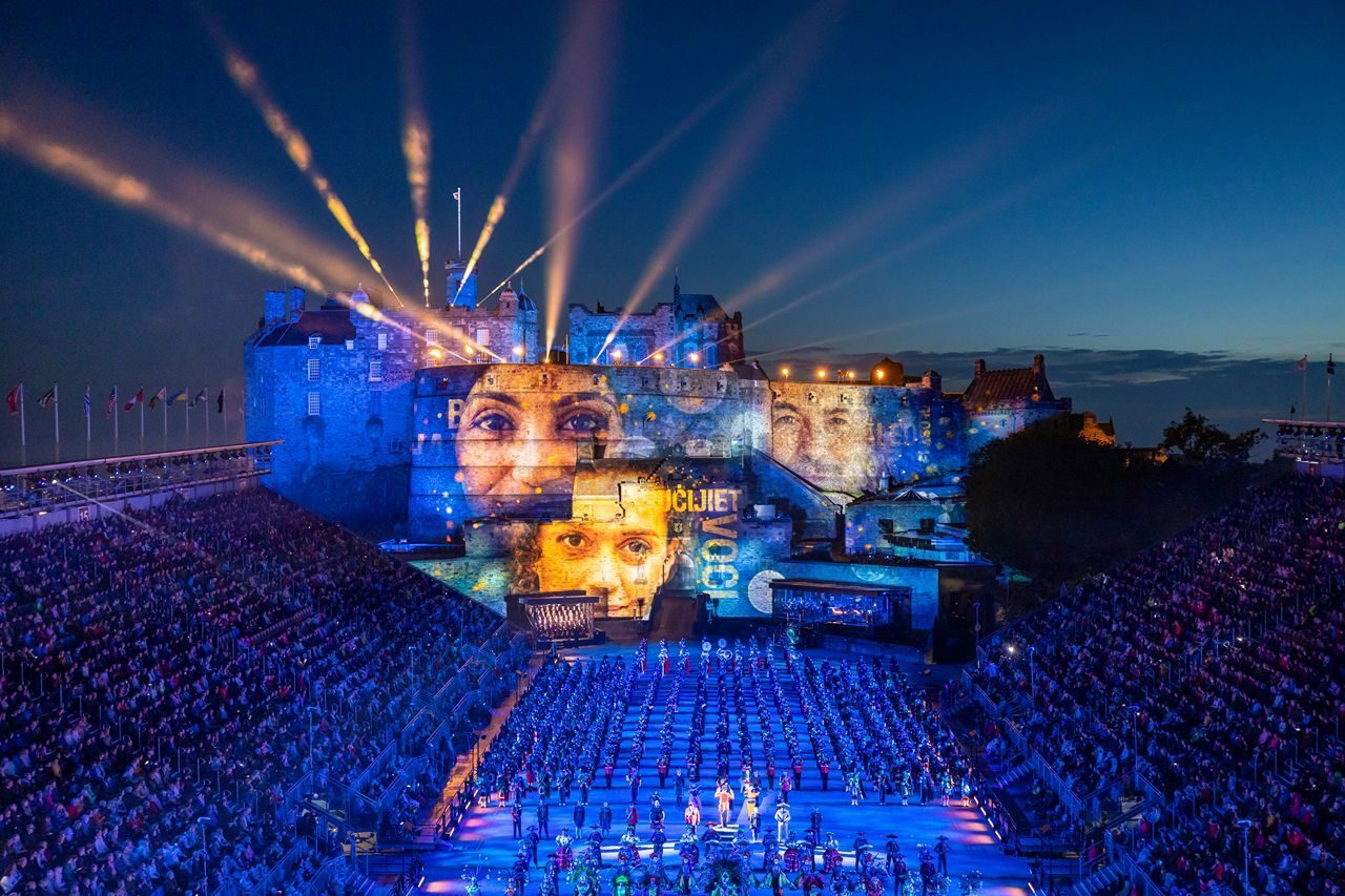 A vibrant night scene of Edinburgh Castle illuminated with projections, showcasing a large audience enjoying a spectacular event.