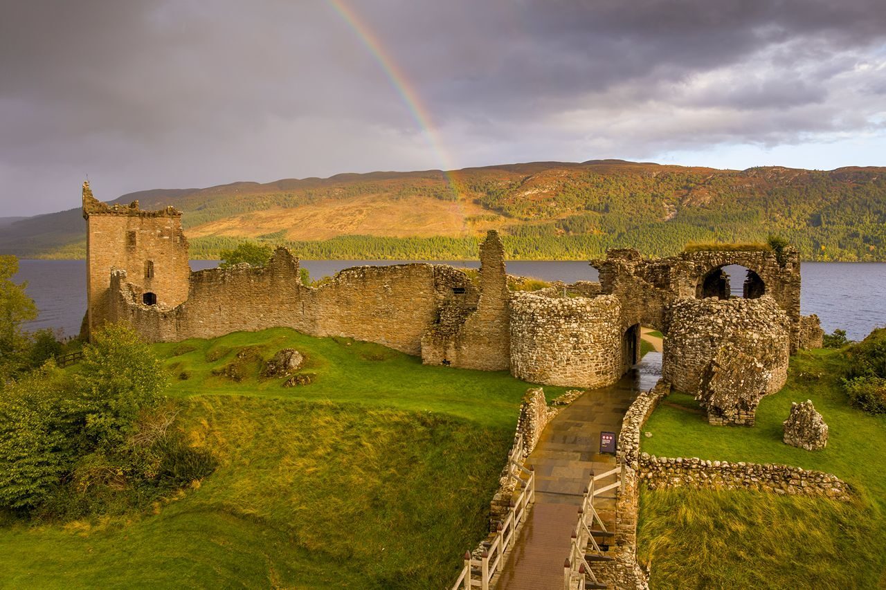 Ruins of a historic castle overlooking a serene lake, with a rainbow arching across a cloudy sky in the background.