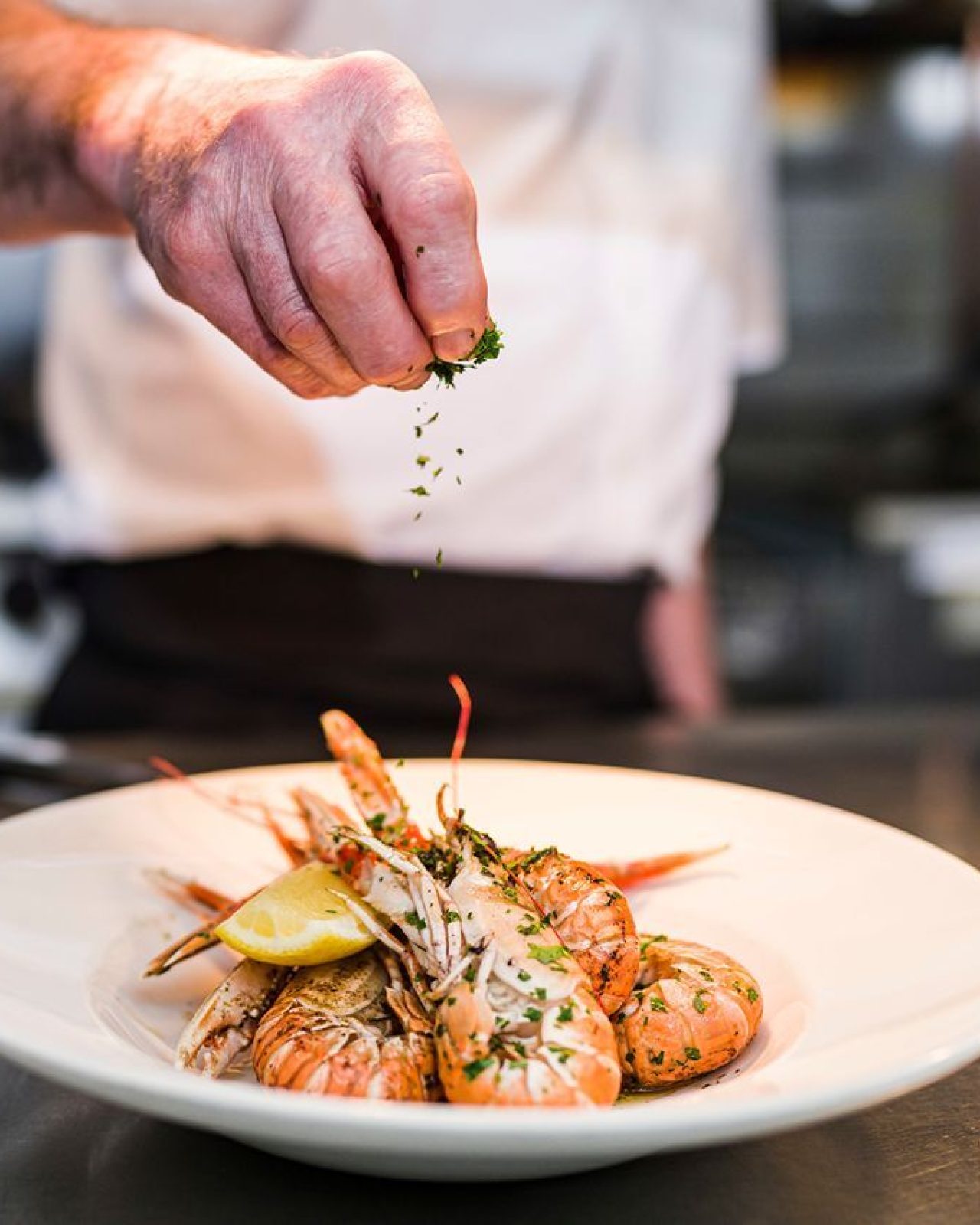 a white plate of langoustine and lemon slice with a chefs hand sprinkling green parsley