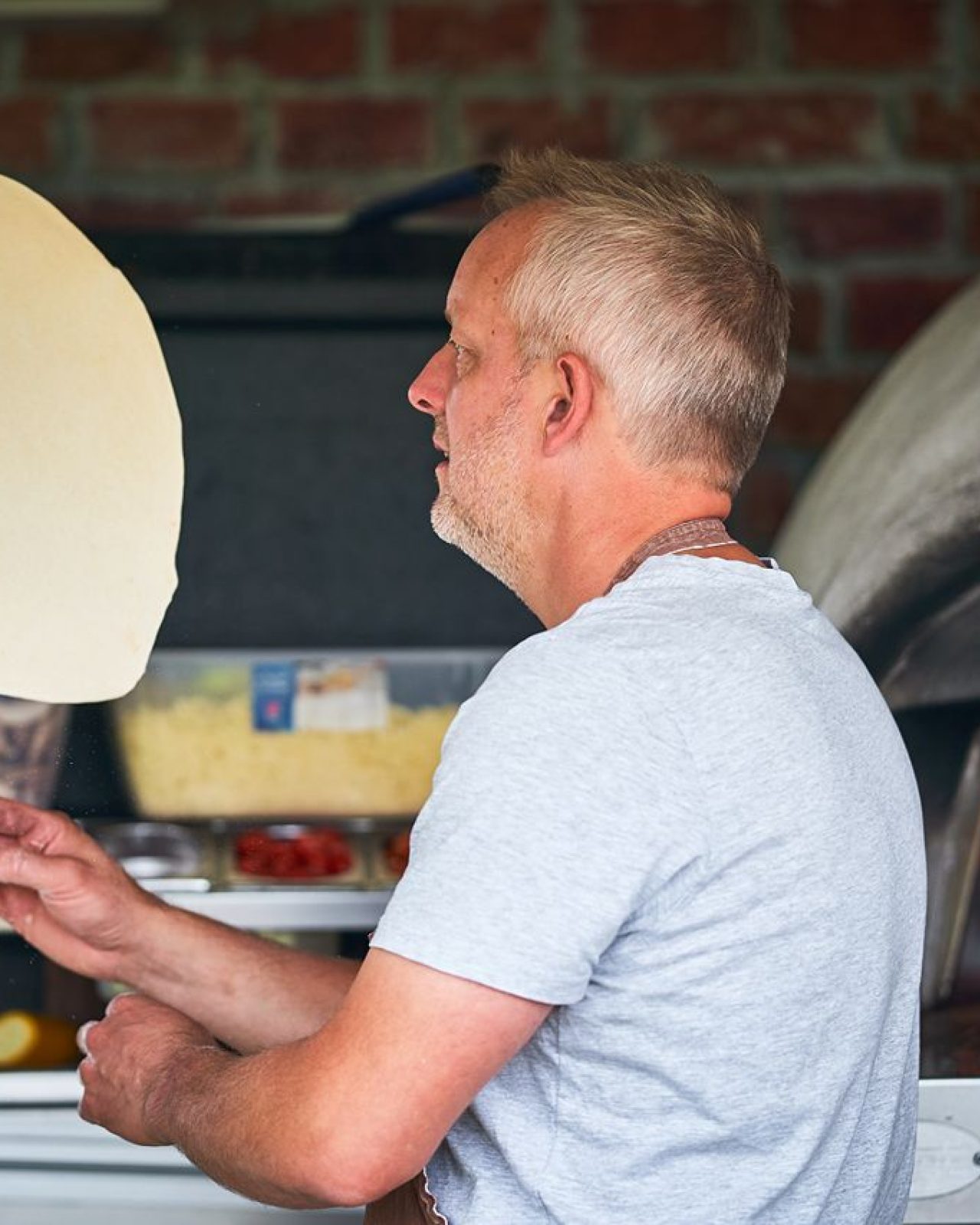 A grey haired man throwing pizza dough standing in front of a wood burning pizza oven