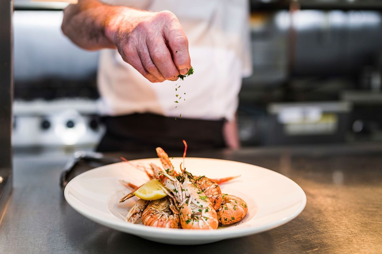 a white plate of langoustine and lemon slice with a chefs hand sprinkling green parsley