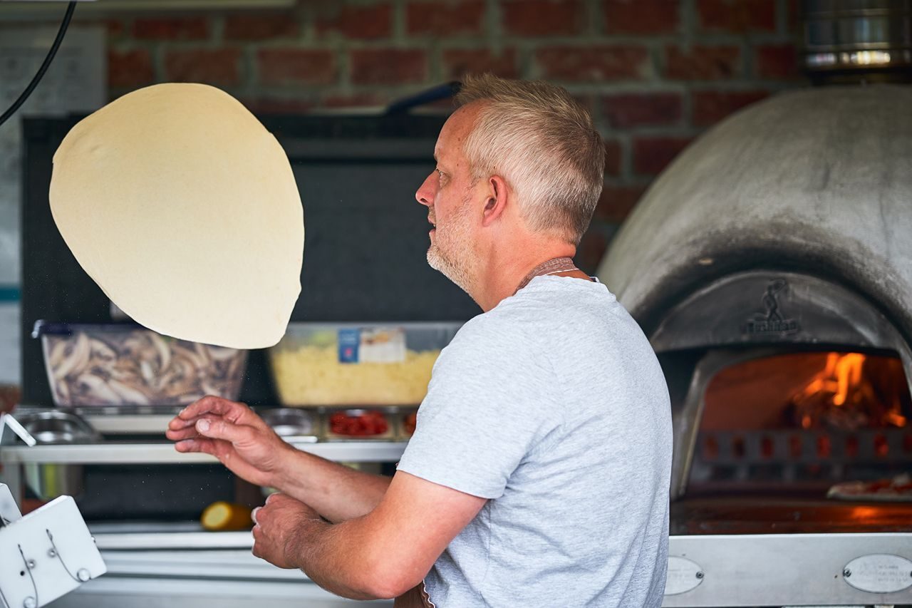 A grey haired man throwing pizza dough standing in front of a wood burning pizza oven