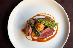 a white, circular plate on a brown wood table with venison, cabbage, potatoes and golden hued jus