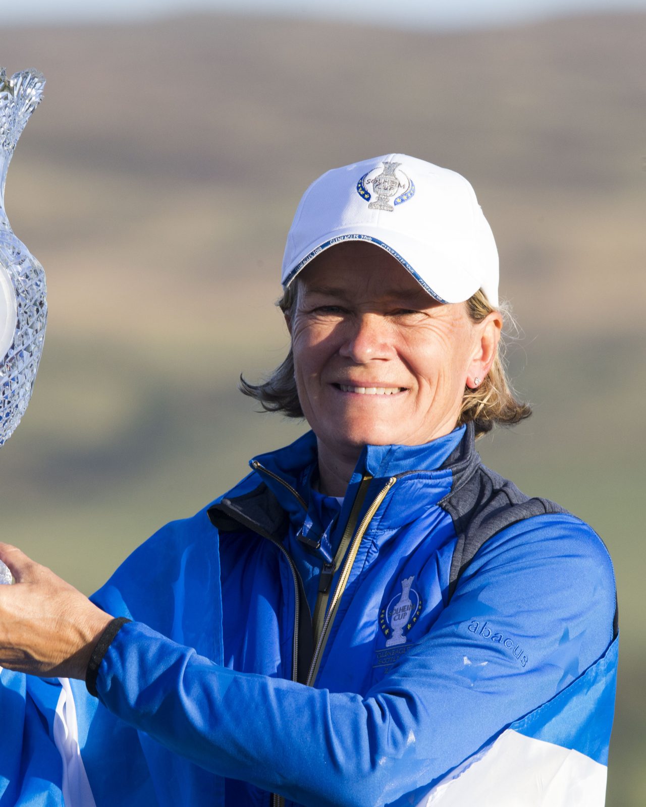 Smiling person in blue and white sports attire holding a large crystal trophy. Sunny outdoor setting with blurred green hills in the background.