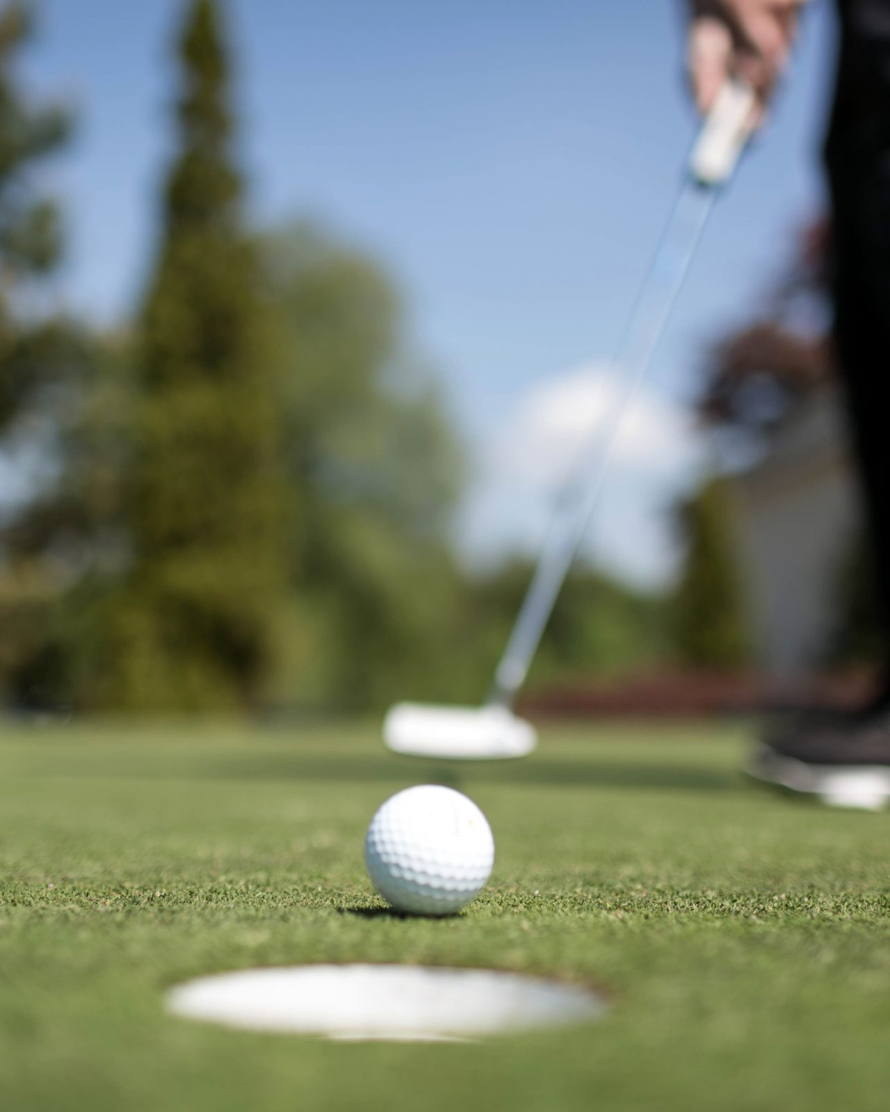 A person prepares to putt a golf ball towards the hole on a putting green, with the ball and hole in focus and the golfer blurred in the background.