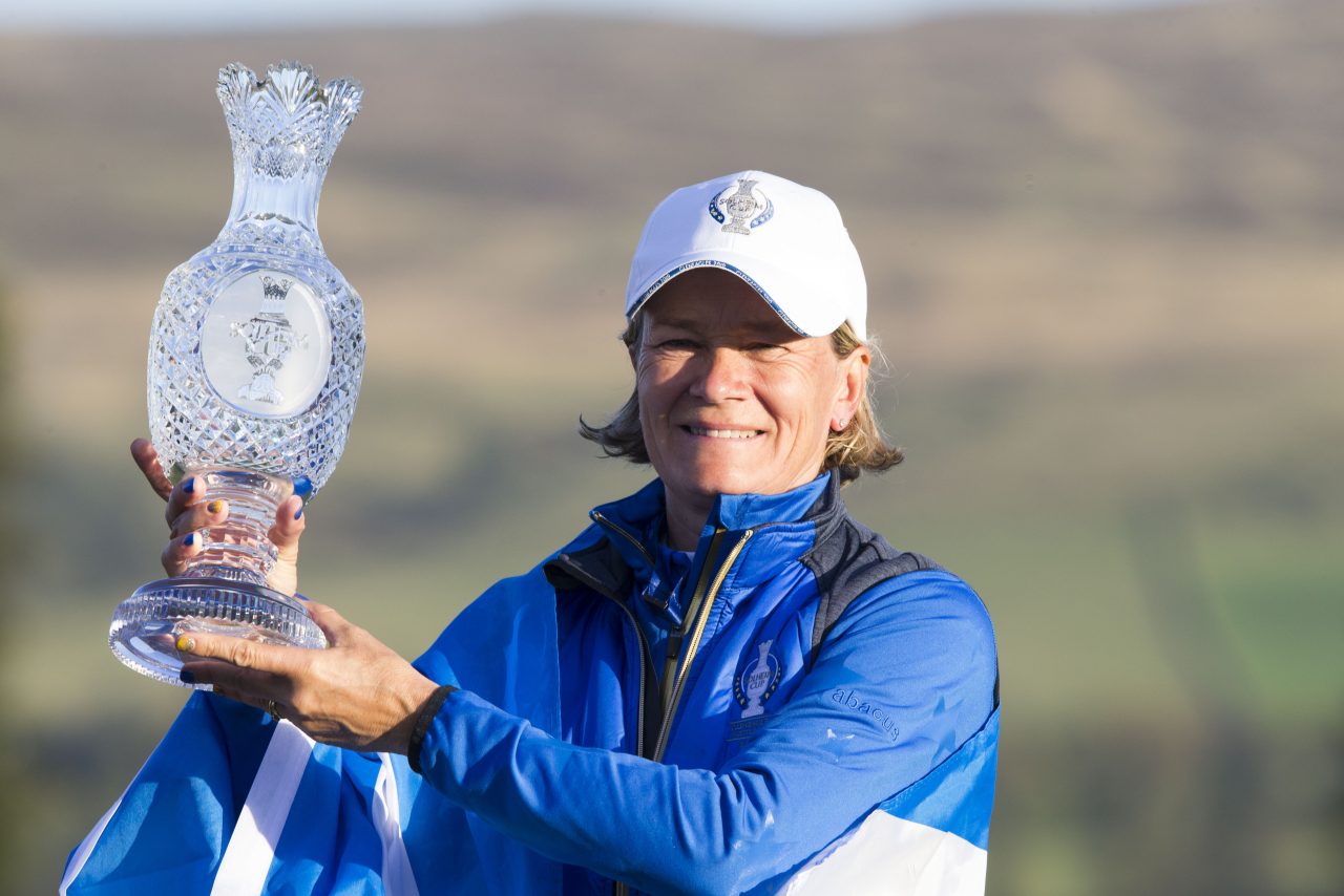 Smiling person in blue and white sports attire holding a large crystal trophy. Sunny outdoor setting with blurred green hills in the background.