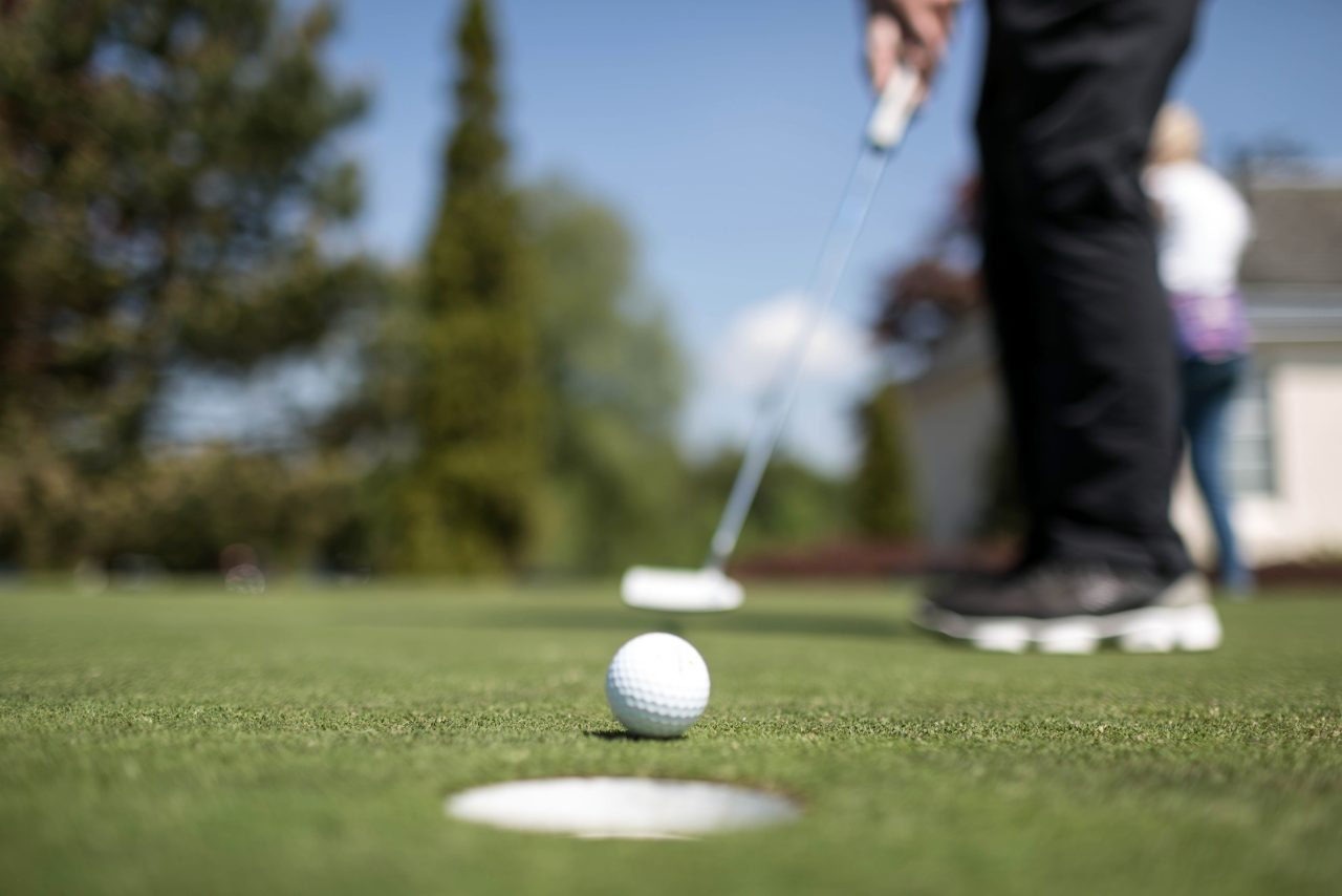 A person prepares to putt a golf ball towards the hole on a putting green, with the ball and hole in focus and the golfer blurred in the background.