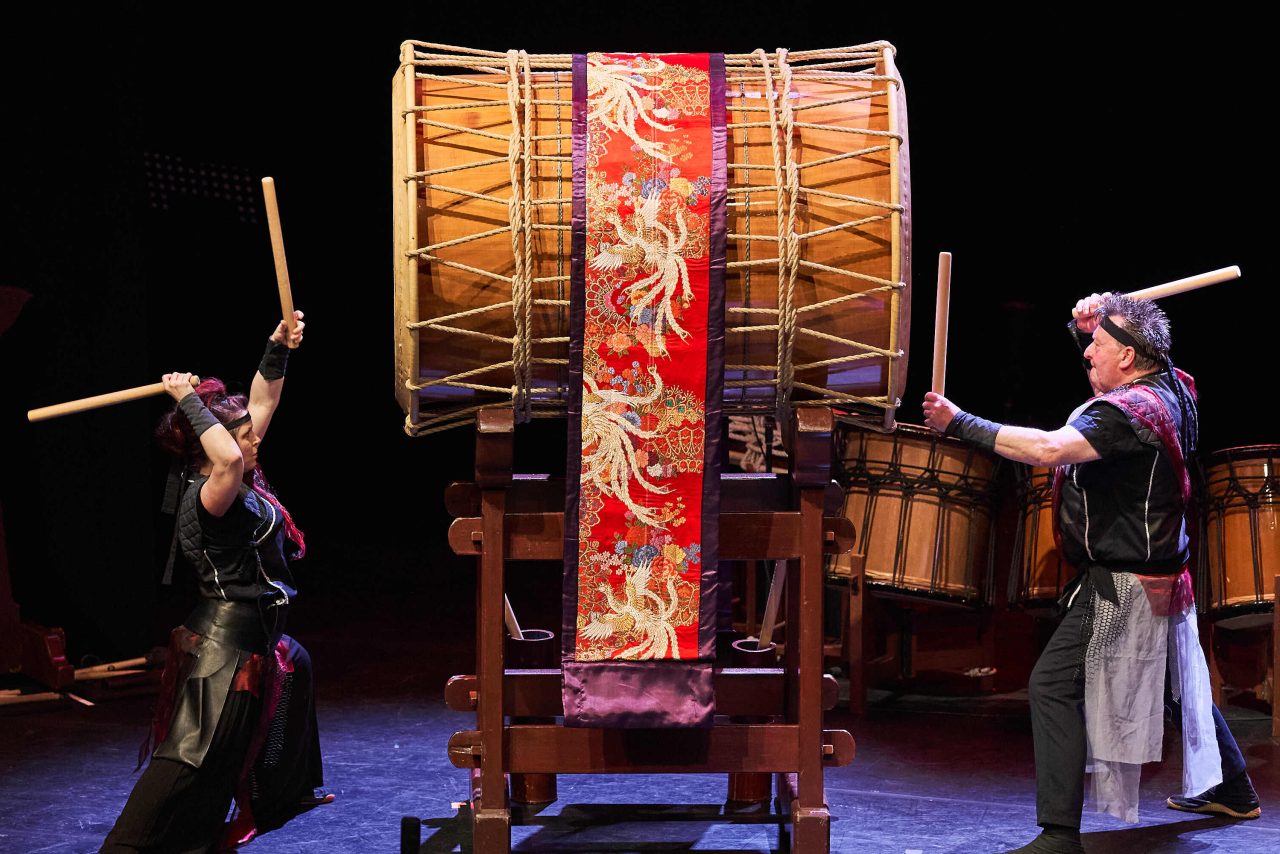 two men, on stage, banging on a giant drum