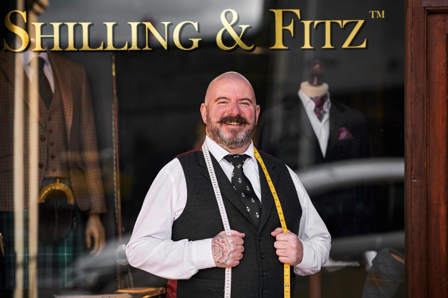 A happy man in a suit with a measuring tape over his shoulders standing outside a building which has 'Shilling & Fritz' written in gold text.