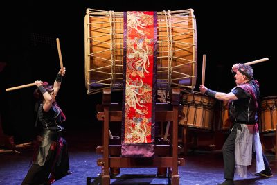 two men, on stage, banging on a giant drum
