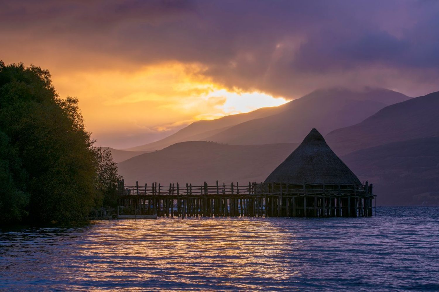 Ancient crannog building reaching into a loch with hills in background, in the early evening light