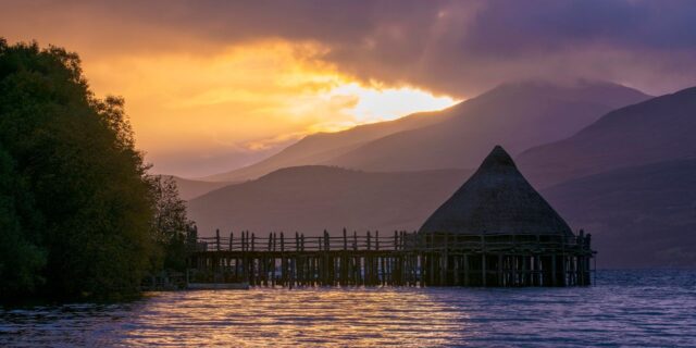 Ancient crannog building reaching into a loch with hills in background, in the early evening light