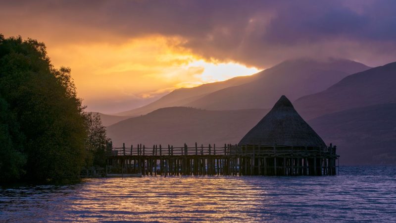 Ancient crannog building reaching into a loch with hills in background, in the early evening light