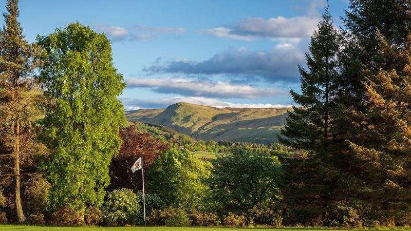 Green golf hole,  area surrounded by large green and red trees on a sunny day with rolling green hills in the distance.