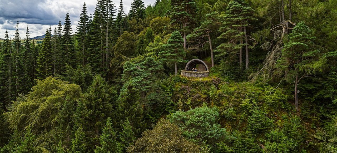 It shows a wooden shelter in the shape of a Pine Cone on top of a hill surrounded by green trees. The image is taken from above.