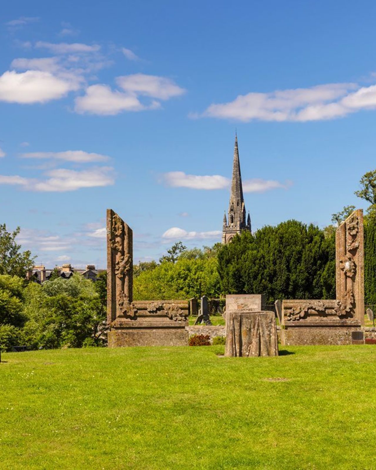 Scenic view of a park featuring historical stone sculptures, lush greenery, and a church steeple against a blue sky.