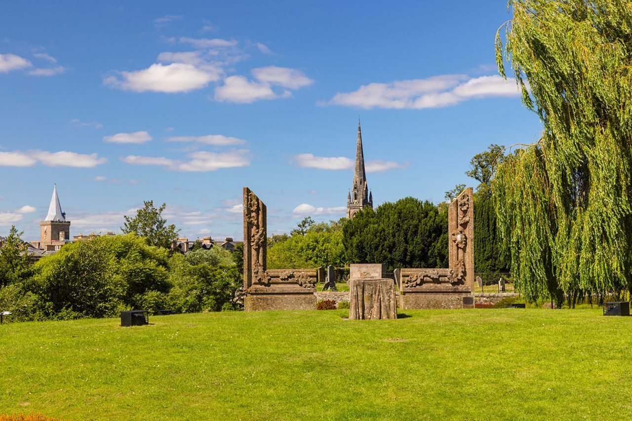 Scenic view of a park featuring historical stone sculptures, lush greenery, and a church steeple against a blue sky.
