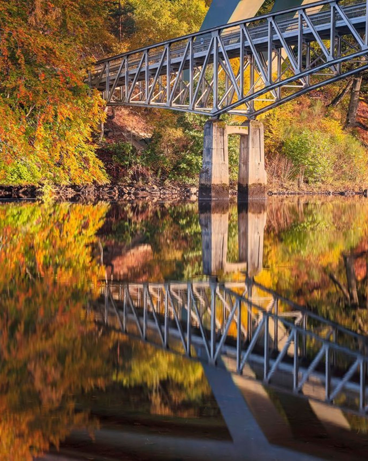 A metal bridge over a calm body of water, reflecting the colourful autumn foliage on the banks.