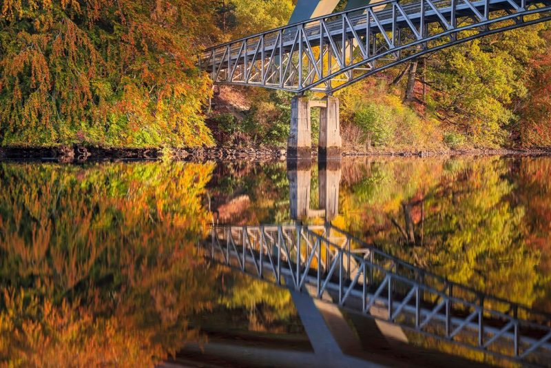 A metal bridge over a calm body of water, reflecting the colourful autumn foliage on the banks.