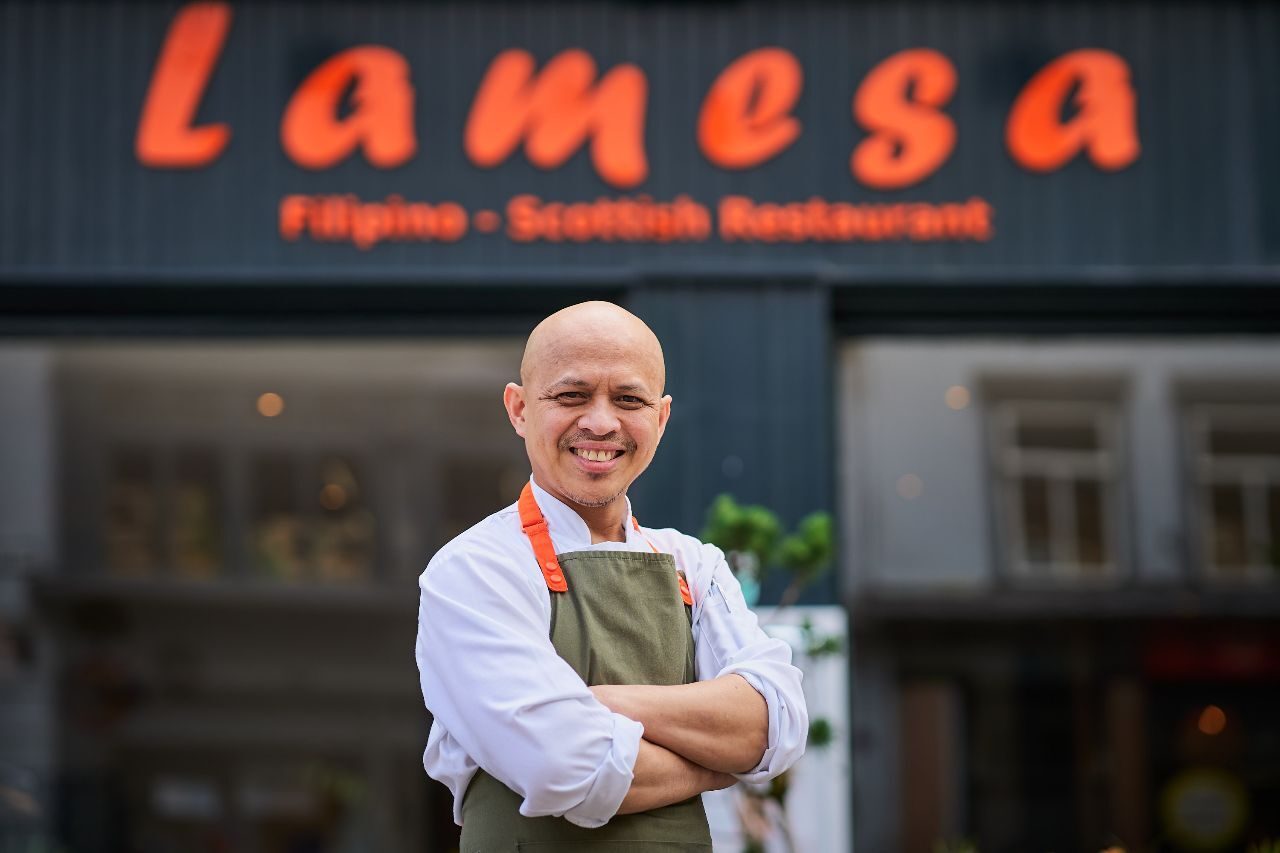 a chef standing outside a black budling with orange signage