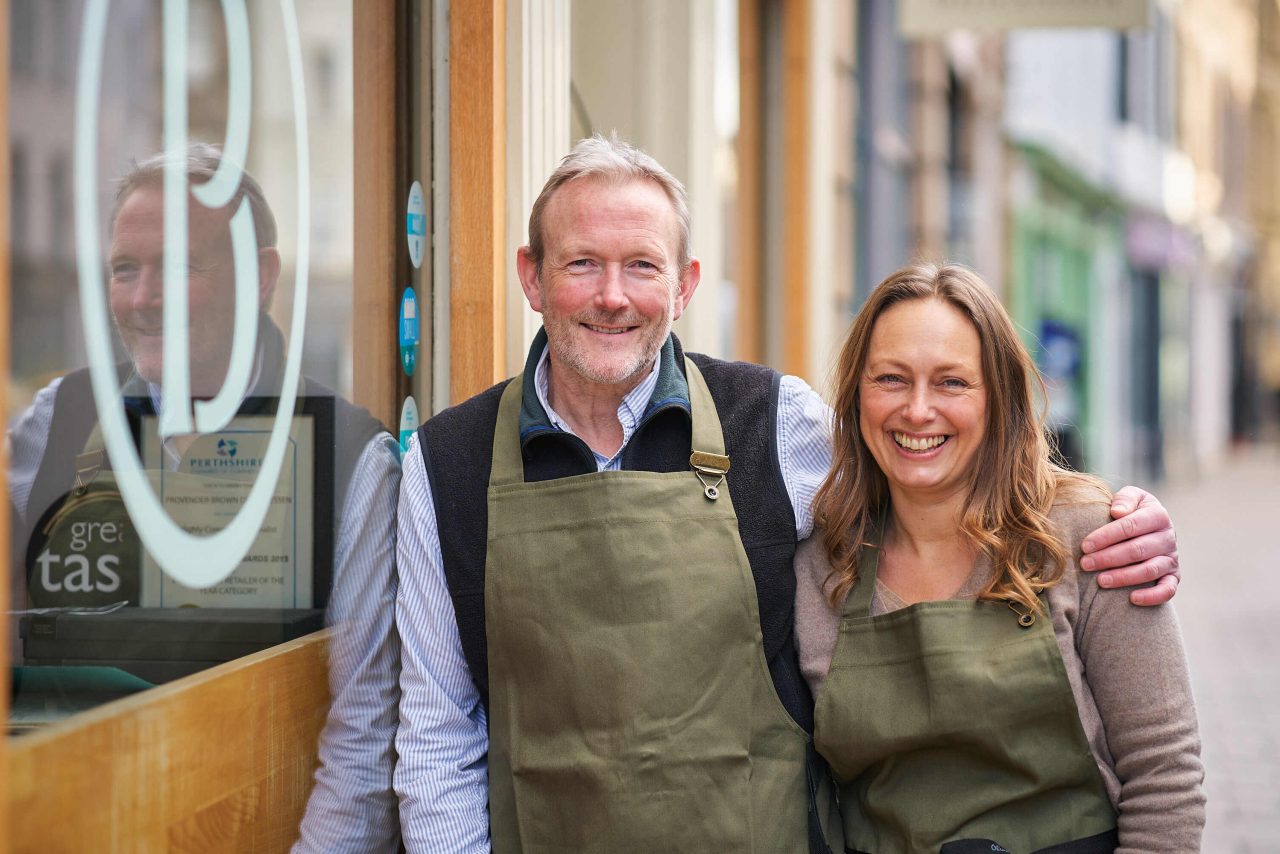 A man wearing an apron rests his hand on the shoulder of a woman wearing an apron