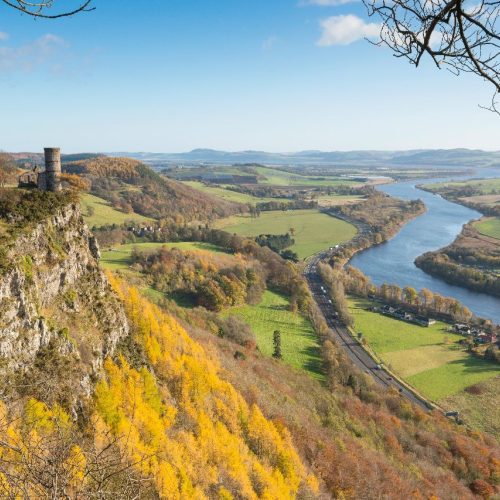 A round stone tower sits at the top of a sheer cliff face above a scenic landscape and the River Tay winding its way to Dundee.