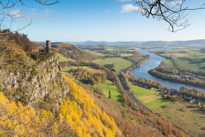 A round stone tower sits at the top of a sheer cliff face above a scenic landscape and the River Tay winding its way to Dundee.