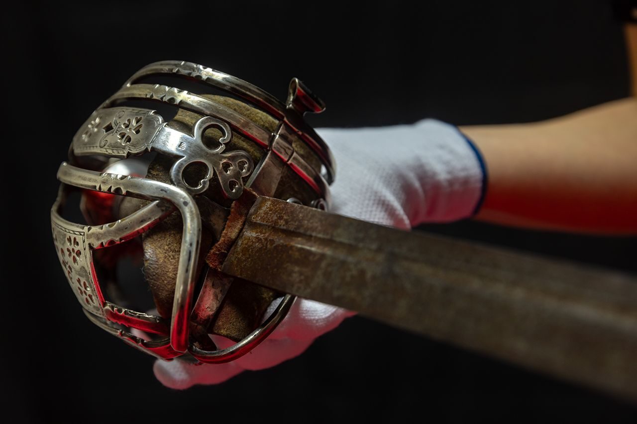 Silver ornate sword handle and blade held by a white gloved hand.