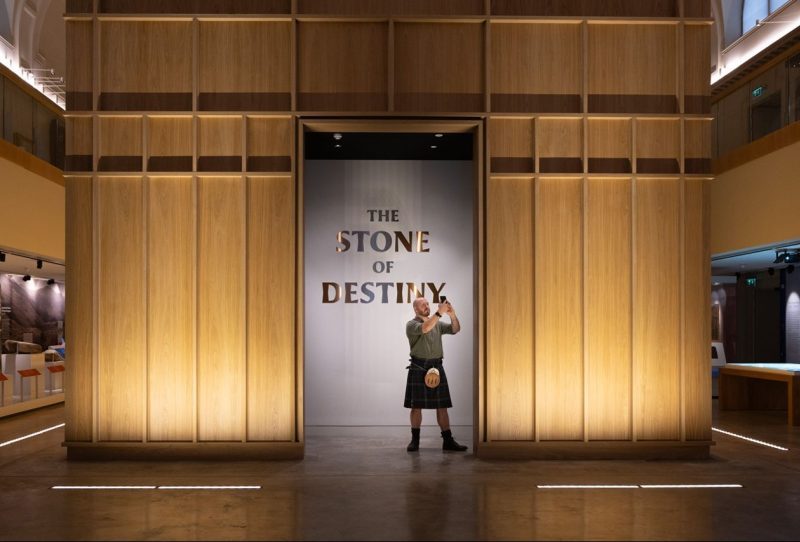 Man in kilt with phone standing in front of Stone of Destiny in Perth Museum