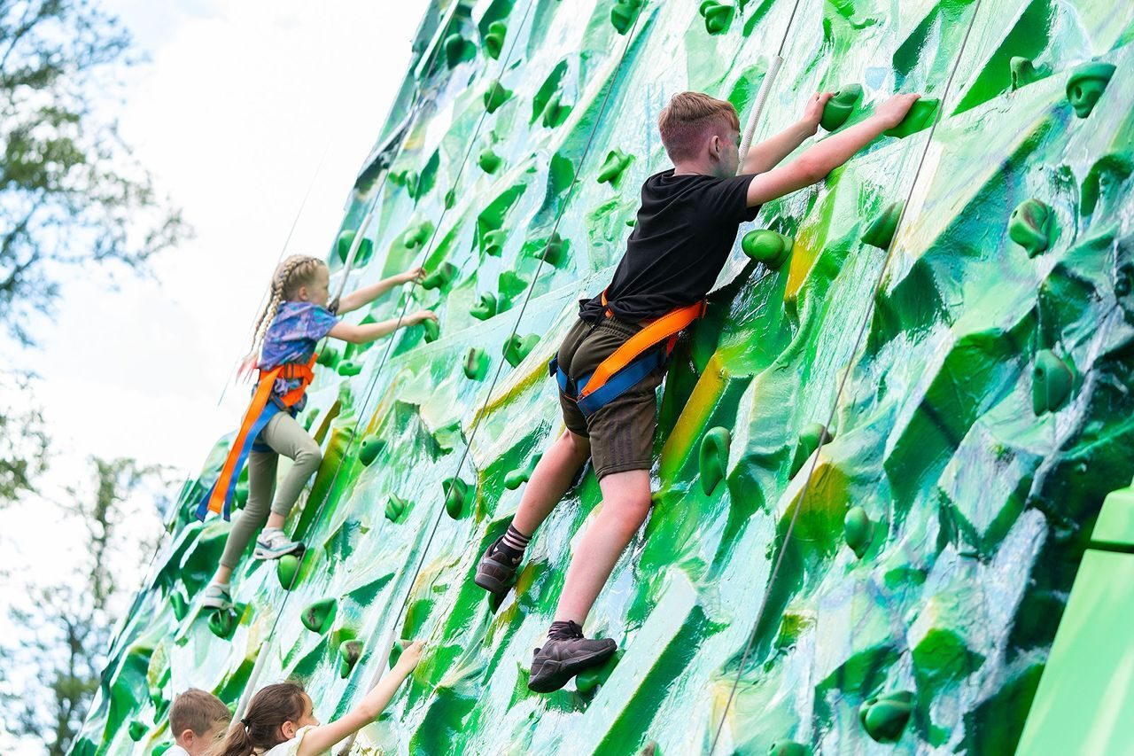 Two children wearing harnesses on a green climbing wall