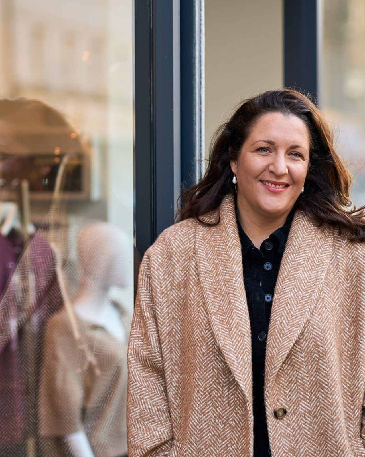 Smiling dark hair woman leaning against a window with items of clothing on a rack behind the glass.