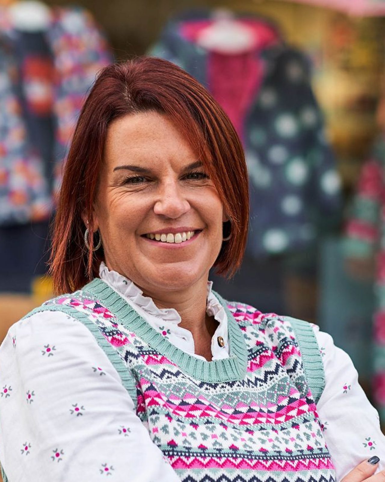 Smiling woman with red hair wearing a pink top in front of a shop window displaying colourful children's clothes.