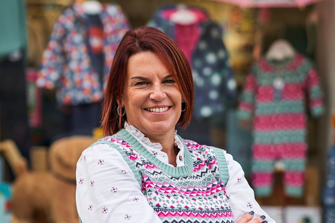 Smiling woman with red hair wearing a pink top in front of a shop window displaying colourful children's clothes.