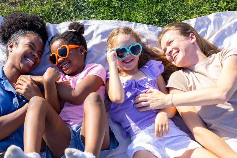 A family lying on a picnic blanket on the grass wearing heart shaped sunglasses and laughing.