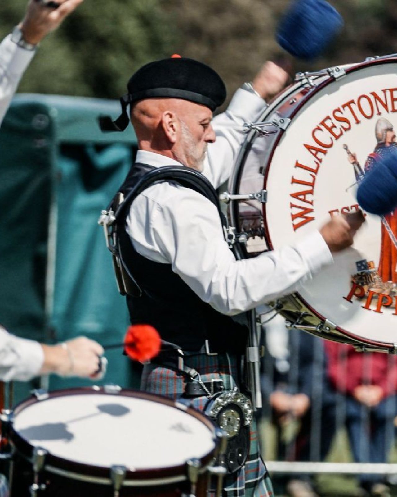 A man wearing a white shirt and green tartan kilt playing a drum which says 'Wallacestone & District Pipe Band.'