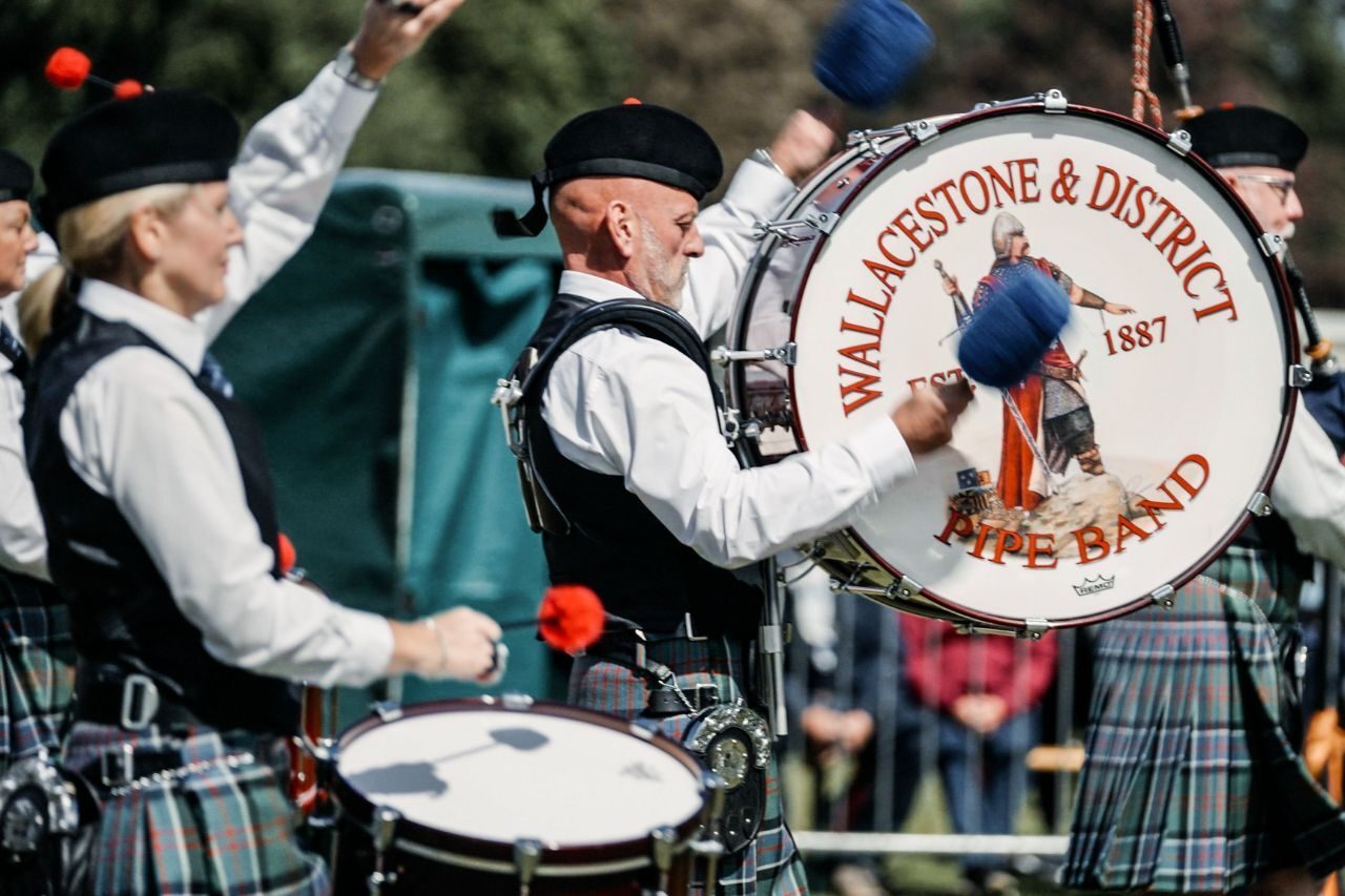 A man wearing a white shirt and green tartan kilt playing a drum which says 'Wallacestone & District Pipe Band.'