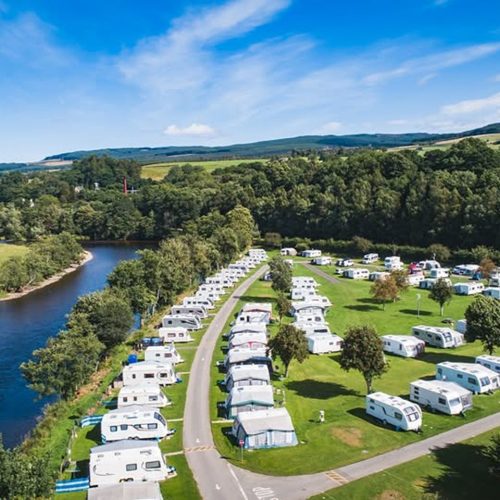 Rows of white static caravans on grass beside a river with trees and hills in the background under blue skies on a sunny day.