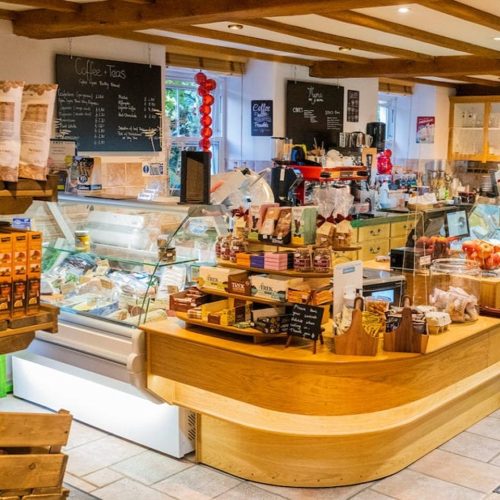 Large wooden counter displaying preservatives, biscuits and other deli goods , beside fridge with cheese and blackboards on walls behind at Thyme deli at Errichel