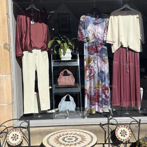 Window displaying three sets of clothing in brown and cream, display unit with bags, table and chairs in foreground.