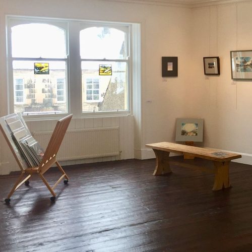 Bright, white room with dark wooden floor displaying art on walls with a wooden bench and large wooden art displayer, with art inside.