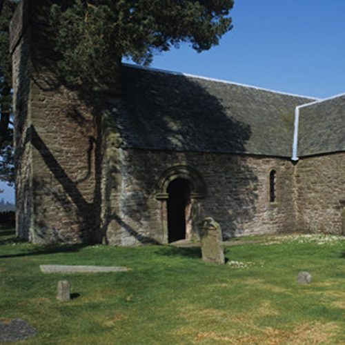Medieval church, with grey old stone in a churchyard with some stone gravestones and a large green tree at lest of picture on a sunny day with blue skies.