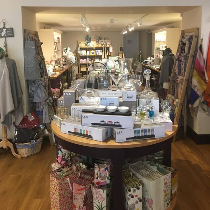 View of a wooden table with gift items on it and underneath it., racks displaying scarves to either  side , large counter at back left of gift shop.