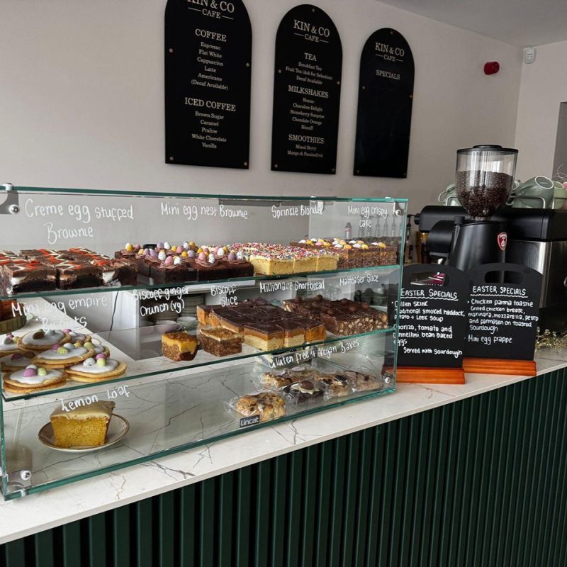 Colourful cakes on display in glass cabinet, on a marble counter with menu boards on back wall for Kin and Co cafe