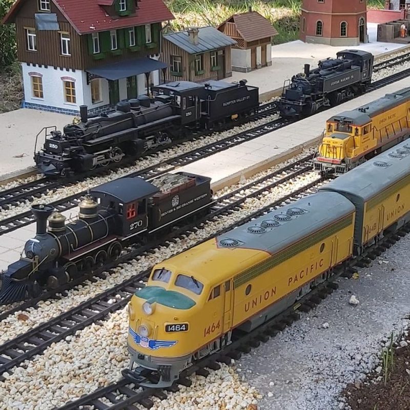 3 bright yellow miniature train, with 3 black trains on tracks beside with a few small station buildings on .